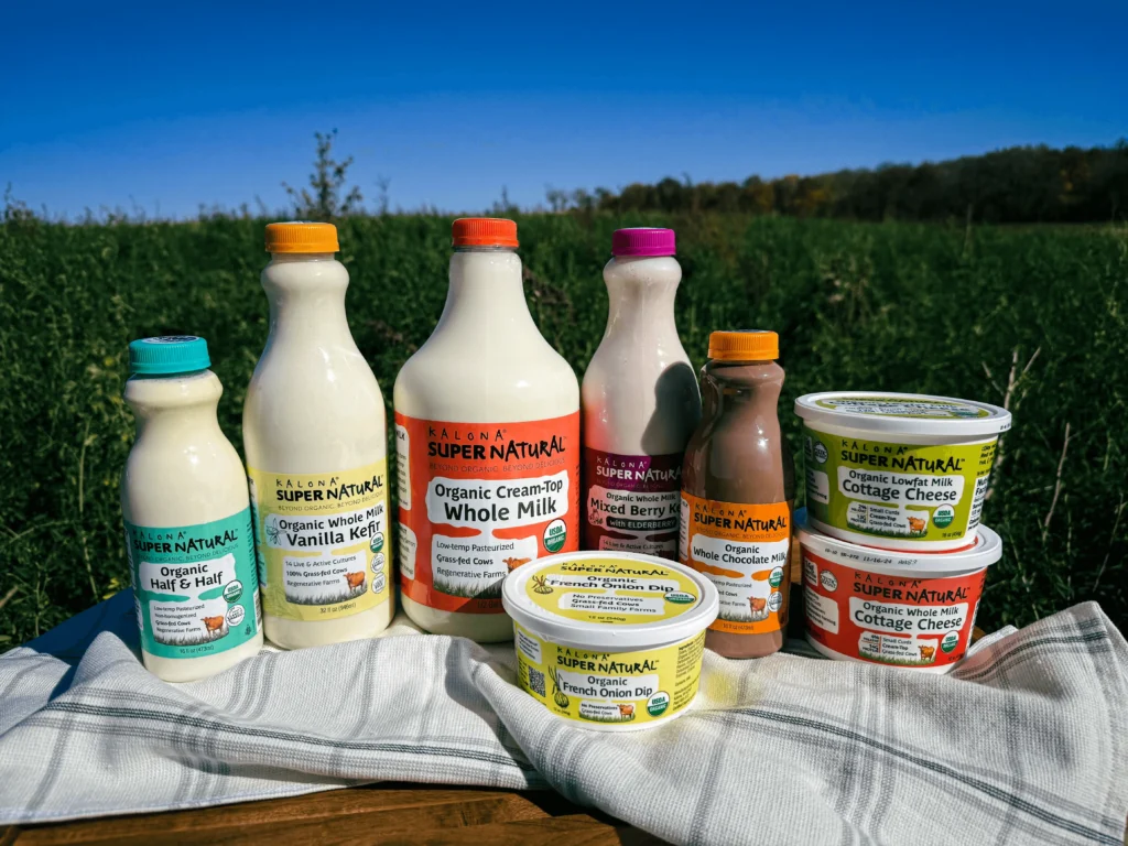 A collection of Kalona SuperNatural organic dairy products, including better milk, kefir, half and half, cottage cheese, and French onion dip, arranged on a white cloth in an open green field under a clear blue sky.